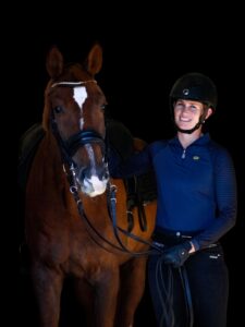 A person in a black riding helmet, blue shirt, and gloves stands next to a brown horse with a white stripe on its forehead, smiling and holding the reins, ready for dressage training against a black background.