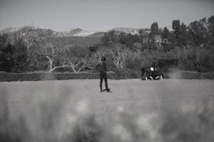 A person stands in the foreground on a sandy equestrian arena, observing dressage training as another rider guides a horse in the background. Trees and distant mountains are visible under a clear sky. The image is in black and white.