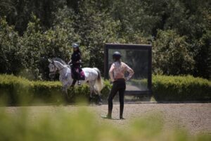 A person on horseback rides past a large mirror outdoors during dressage training, while another person, wearing a helmet and riding gear, stands nearby observing. Greenery and trees surround the arena.