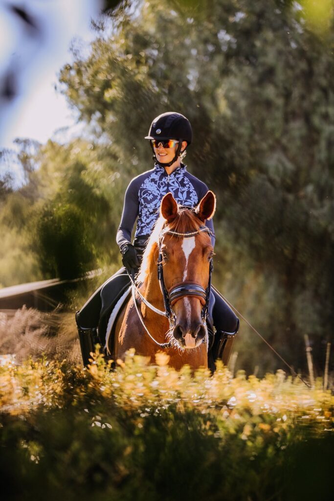 A person wearing a helmet and sunglasses rides a brown horse outdoors, surrounded by greenery and sunlight filtering through the trees.