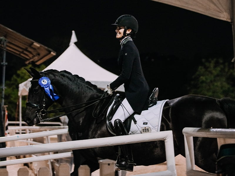 A rider in formal equestrian attire sits on a black horse adorned with a blue ribbon, indicating a win. They are in an outdoor, covered arena at night, with tents and fences visible in the background.