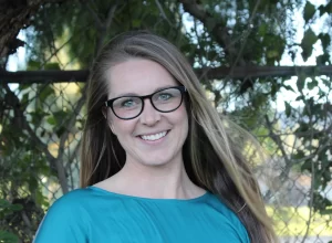 A woman with long brown hair and glasses smiles at the camera. She is wearing a teal top and standing outdoors in front of a chain-link fence and leafy trees, looking ready for her Rider Position Masterclass.