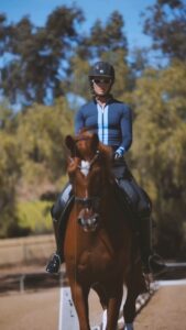 A person wearing a helmet and blue riding outfit is riding a brown horse outdoors, with trees and blue sky in the background.