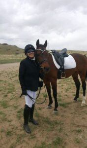 A person wearing equestrian gear stands smiling next to a brown horse with a white saddle pad in an open, grassy field under a cloudy sky, celebrating the spirit of giving back this Black Friday 2022.