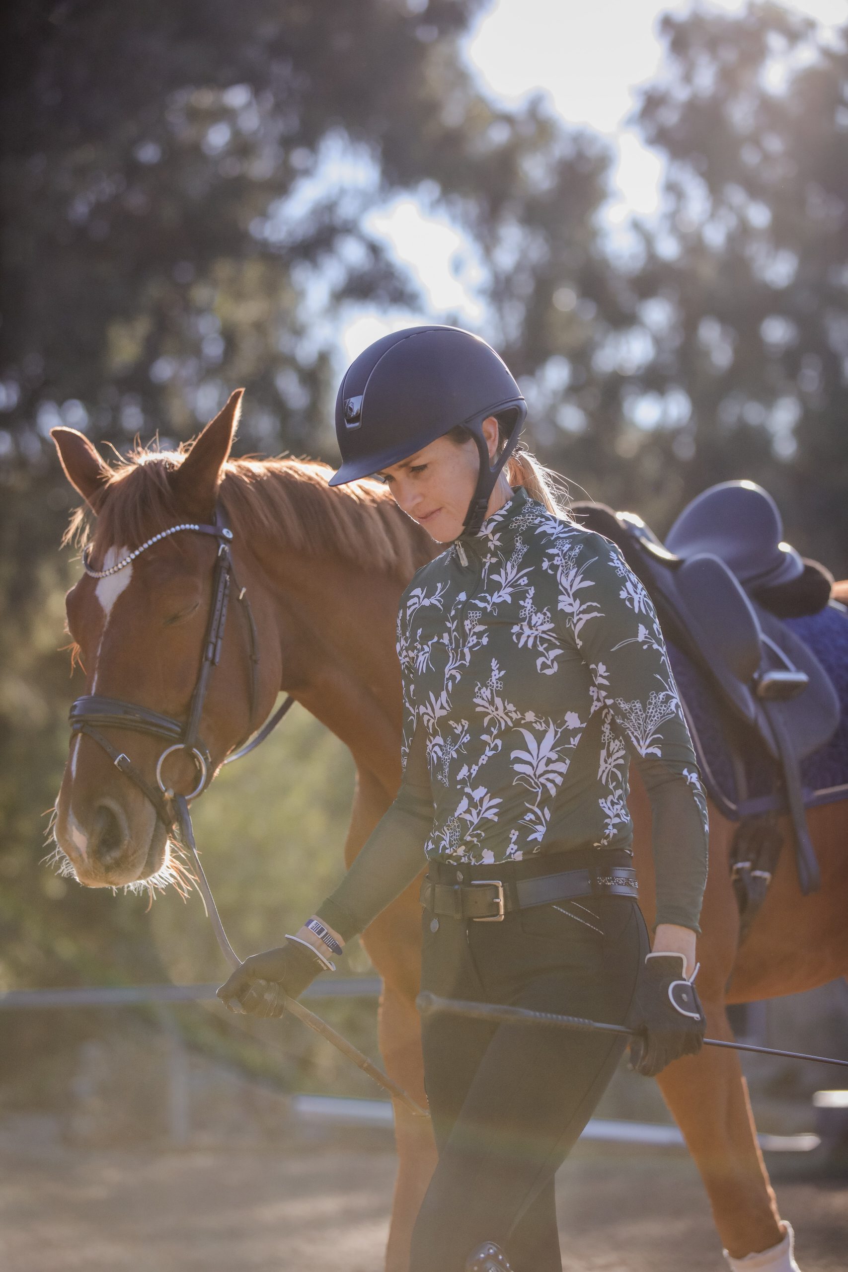 A woman wearing a helmet and floral-patterned shirt leads a brown horse by the reins outdoors on a sunny day, practicing techniques from a Groundwork Masterclass, with trees and sunlight in the background.