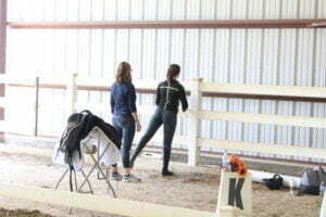 Two women stand in an indoor riding arena near a white fence, discussing Rider Fitness. One points ahead while the other listens. A saddle and tack rest on a stand nearby, and a letter K marker with orange flowers is visible in the foreground.