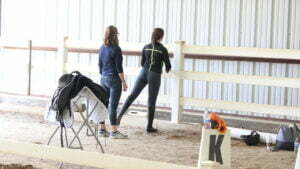 Two women stand in an indoor riding arena next to a white fence, with one resting her foot on the rail—perhaps discussing their Rider Fitness. Nearby, a saddle sits on a rack and a marker with the letter K and a small vase of flowers are visible.