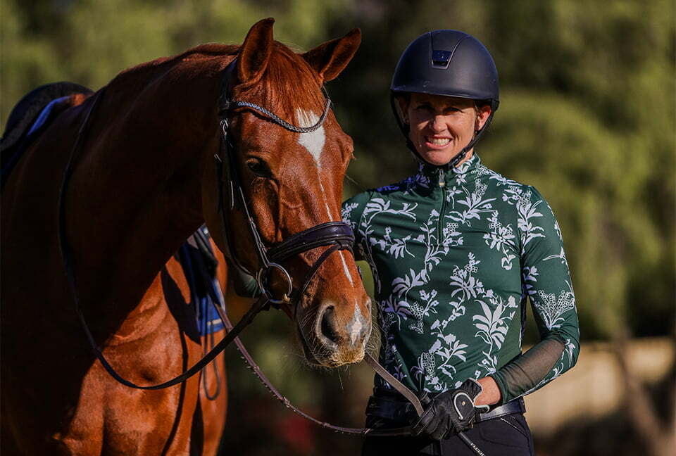 A smiling person in a green floral shirt and black helmet stands beside a brown horse, gently holding its bridle. They are outdoors amidst greenery, ready for groundwork training in a serene setting.