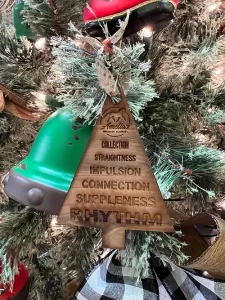 A wooden Christmas tree ornament engraved with equestrian terms hangs on a pine tree, celebrating the Holiday Season 2022. The words COLLECTION, STRAIGHTNESS, IMPULSION, and more are featured, with other ornaments nearby.