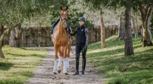 A person in riding gear demonstrates excellent rider position on a tree-lined path, smiling and holding the reins of a brown horse wearing a saddle and white leg wraps. Sunlight filters through the trees onto the green grass in this 2023 scene.