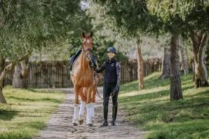 A person in riding gear stands on a tree-lined path, smiling and holding the reins of a saddled brown horse with white leg wraps. Sunlight filters through the trees as their 2023 rider position creates a serene outdoor scene.