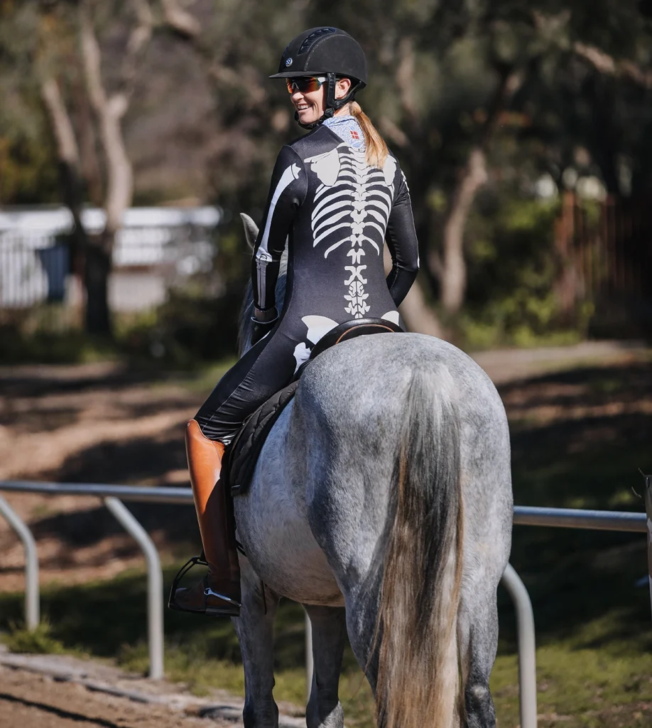 A person wearing a black bodysuit with a white skeleton print and a helmet is riding a gray horse outdoors, smiling and looking back over their shoulder, showcasing their Rider Ranking for 2023.