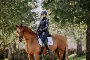 A smiling person in equestrian gear showcases excellent rider position while sitting on a brown horse outdoors, surrounded by green trees and sunlight filtering through the branches in 2023.