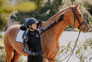 A smiling rider in a 2023-patterned jacket and helmet stands beside a chestnut horse, gently touching its neck. The horse wears a bridle and saddle pad, and their rider position is relaxed outdoors near water and greenery.