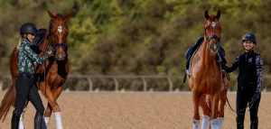 Two women in riding helmets and equestrian gear stand outdoors, each holding the reins of a brown horse. The scene is set on a sandy arena with a blurred green background—perfect inspiration for your next horse personality quiz.