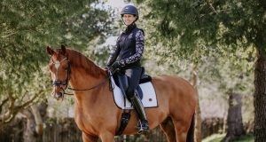 A person wearing riding gear and a helmet sits smiling on a brown horse with a white Custom Saddlery saddle pad, surrounded by green trees in an outdoor setting—perfect for an interview about their equestrian journey.