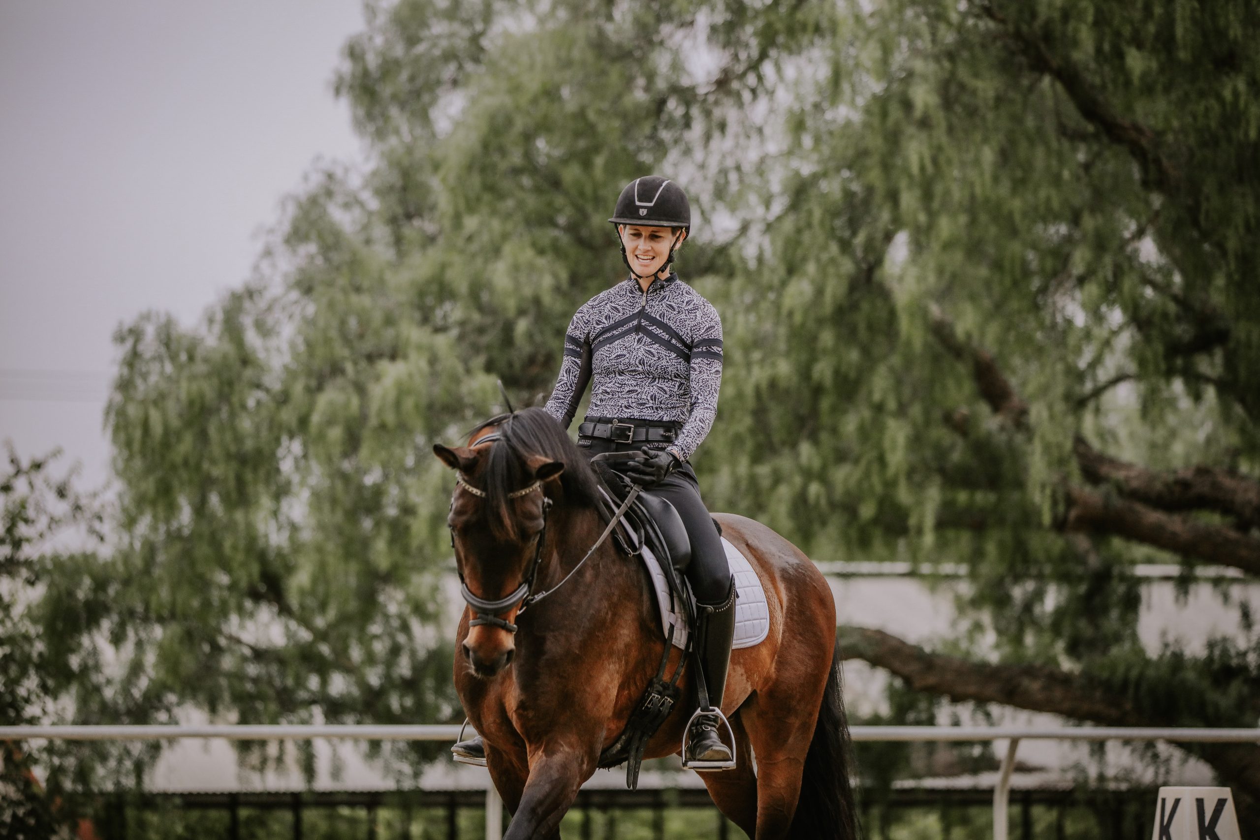 A person in equestrian gear, including a helmet and patterned long-sleeve shirt, rides a dark brown horse in an outdoor arena with trees in the background, as seen during a July 2023 event featured in the news.