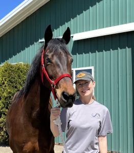 Strides Member Cindi, wearing a gray t-shirt and cap, smiles next to a brown horse in a red halter. They stand outside a green metal building on a sunny day.