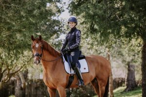 A person wearing a helmet, black riding vest, and boots sits on a brown horse with a white saddle pad, outdoors among green trees and sunlight—a scene reminiscent of August 2023 News highlights.