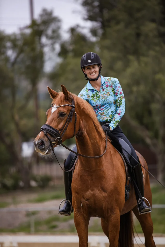 A smiling equestrian, taking part in the 30 Days Challenge, wears a floral-patterned shirt and black helmet while sitting on a chestnut horse outdoors, with trees in the background.