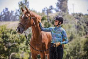 A person wearing a helmet and floral shirt smiles while standing beside a brown horse, holding its lead and gently touching its neck—a perfect moment showcasing horse training and the importance of good ground manners on a sunny day.