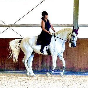 Carol Anne Marano, wearing a black helmet and outfit, rides a white horse indoors. The horse sports protective leg gear and a maroon ear bonnet as sunlight filters into the arena with light sand on the ground.