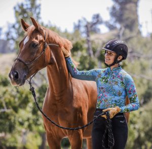 A woman in a helmet and floral top smiles while standing beside a chestnut horse, gently petting its neck outdoors on a sunny day. Trees and greenery are visible in the background, evoking the joy of connecting with horses and exploring free resources for riders.