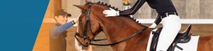 A person on horseback receives guidance from a smiling trainer during dressage training. The horse has a braided mane and wears English riding tack. The indoor scene features a blurred background and an orange graphic on the left.