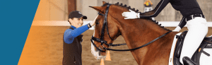 A smiling instructor in a cap and vest gently pats a brown horse’s face while communicating with its rider, who is dressed in black and white riding gear, during dressage training inside an indoor arena.