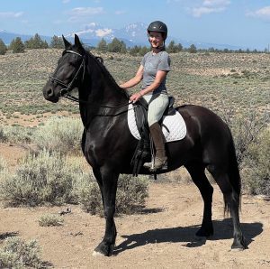 Strides member Rebekah, wearing a helmet and riding gear, sits on a black horse with a white saddle pad outdoors in a dry, shrub-dotted landscape with mountains and blue sky in the background.
