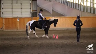 A person on a black and white horse, nicknamed Snowman, is inside an indoor arena approaching orange cones, while another observes—perfect for teaching riding skills. The arena has wooden and metal walls and a dirt floor.