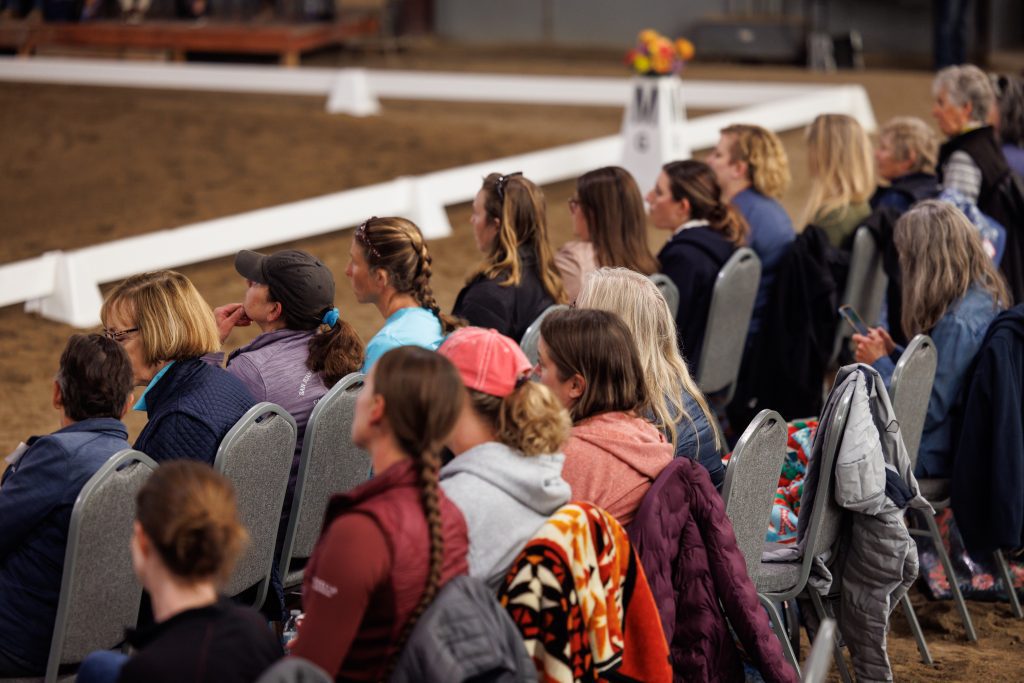 A group of people sit in rows of chairs at an indoor arena, facing away from the camera and watching an event. Some wear hats and jackets, and there is a white fence and flowers in the background.
