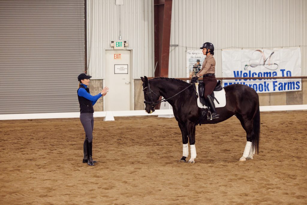 A horse rider in a helmet sits on a black horse inside an indoor arena, while an instructor in riding gear stands nearby, gesturing with both hands. A Welcome to Millbrook Farms banner hangs in the background.
