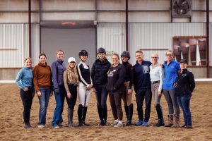 A group of thirteen people, some wearing equestrian helmets and riding attire, stand side by side smiling in an indoor riding arena with sandy flooring and metal walls.