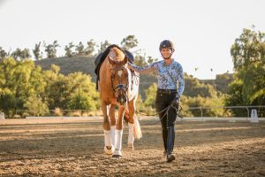 Amelia Newcomb smiling and leading a chestnut horse at a walk.