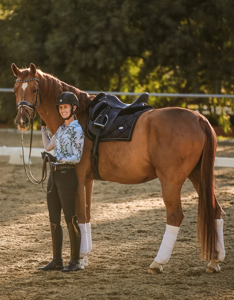 A person wearing a helmet, floral shirt, and riding boots stands smiling next to a saddled chestnut horse with white leg wraps in an outdoor riding arena, ready to make productive Strides, with trees in the background.