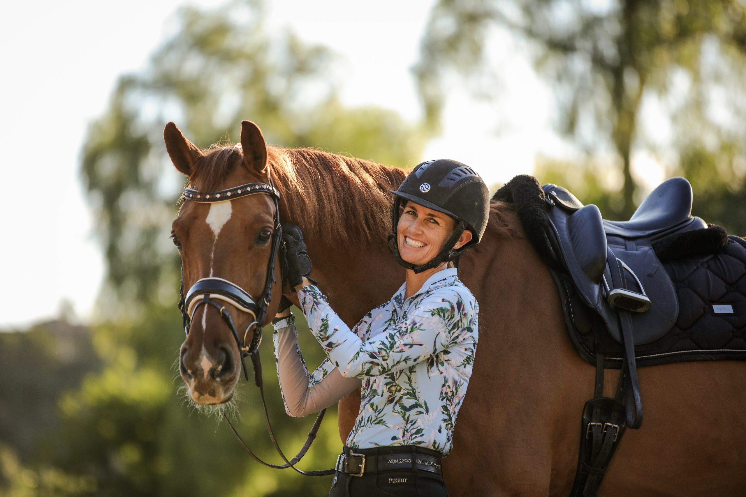 Amelia Newcomb smiling and posing with her chestnut horse who is wearing a dressage saddle and snaffle bridle.