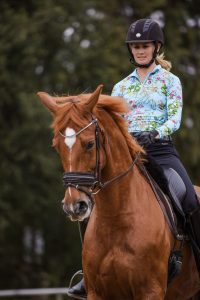 A woman wearing a helmet and a floral-patterned shirt rides a brown horse with a white marking on its forehead, embodying personal growth against a backdrop of blurred green foliage.