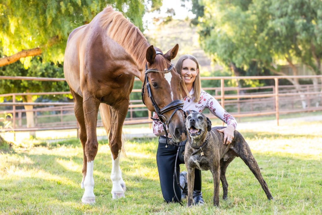 A woman kneels on grass, smiling beside a brown horse and a brindle dog in a sunny, fenced outdoor area with trees behind them—capturing the joy and calm after a mindset workshop.