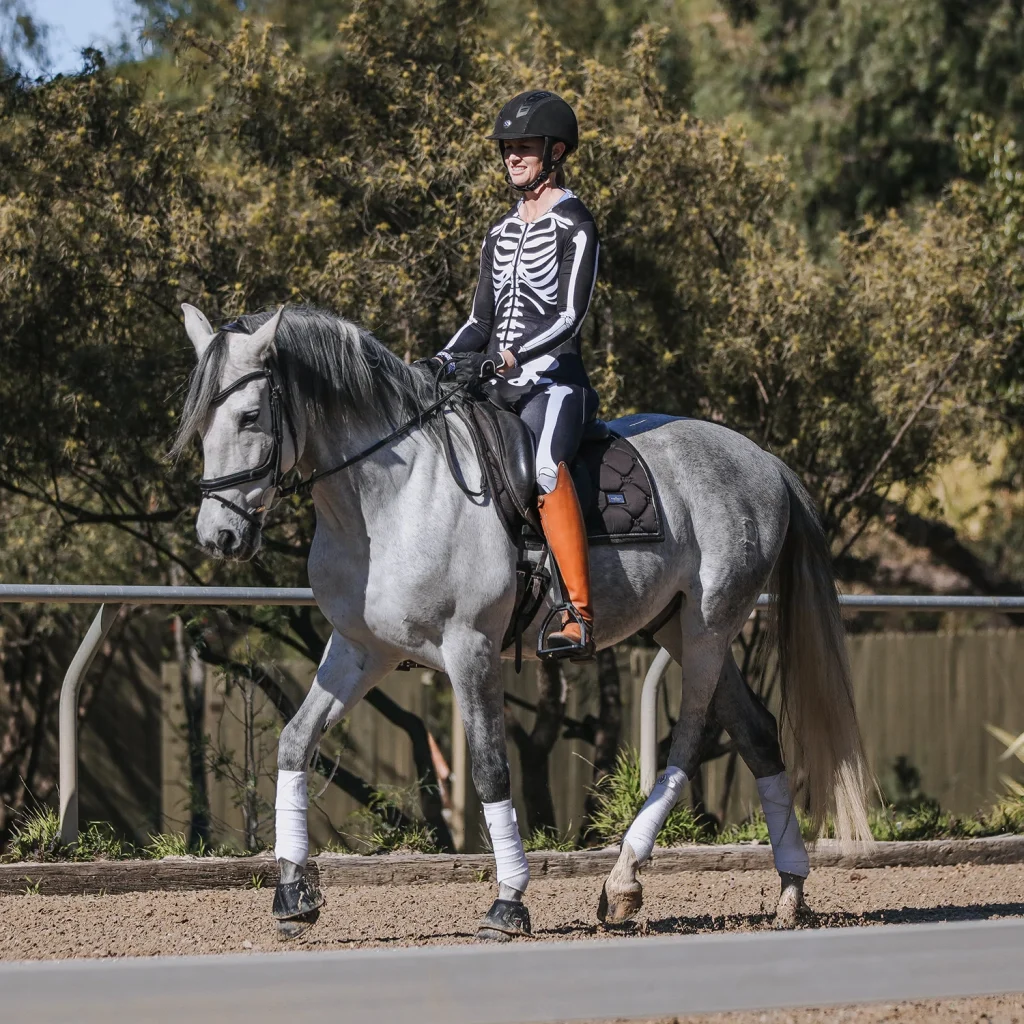 A person wearing an equestrian helmet and a skeleton costume takes on the Sitting Trot Challenge, riding a grey horse with white leg wraps and orange boots along an outdoor path, surrounded by trees in the background.