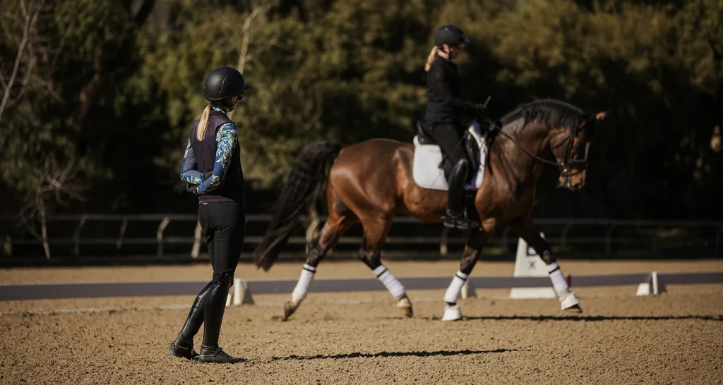 A person on horseback practices dressage in an outdoor arena, focusing on the Sitting Trot Challenge, while another rider in helmet observes from the ground. Trees and sunlight create a scenic backdrop.