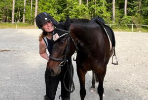 A person wearing a helmet and riding gear stands next to a dark brown horse on a gravel surface, green trees behind them. Smiling, they lean close to the horse’s head, ready to take on the Sitting Trot Challenge together.