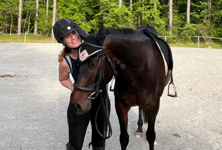 A person wearing a helmet and riding gear stands next to a dark brown horse on a gravel surface, green trees behind them. Smiling, they lean close to the horse’s head, ready to take on the Sitting Trot Challenge together.
