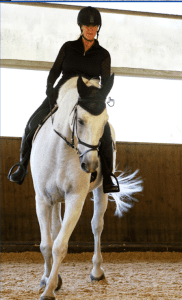 A person wearing a helmet and black riding attire is riding a white horse indoors on a sandy surface, likely engaged in competition training, with wooden walls and large windows in the background.