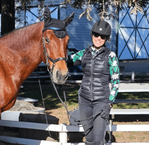 A smiling woman in riding gear stands next to a brown horse wearing a black fly bonnet, holding its reins. They are outdoors near a white fence, with trees and a blue building—capturing the spirit of the 5 Day Riding Bootcamp.