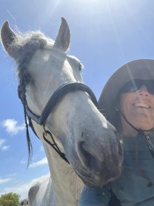 A person in a wide-brimmed hat and sunglasses smiles next to a white horse with a braided mane under a sunny, blue sky at the 2024 Bootcamp.