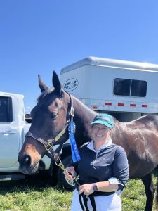 A smiling woman in a green cap and white pants stands next to a brown horse with a blue ribbon, after a 5 Day Riding Bootcamp, with a horse trailer and white truck in the background on a sunny day.