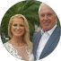 A smiling woman in a white dress stands next to a smiling man in a blue suit, perhaps celebrating after competition coaching. Green palm tree leaves are visible in the blurred background.