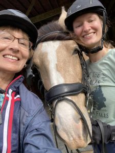 Two smiling women in helmets pose for a selfie with a light brown horse at the 5 Day Riding Bootcamp. The horse wears a bridle, and the women—one older, one younger—look happy and close together during this special riding bootcamp experience.