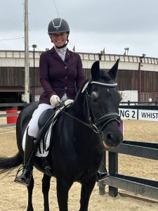 A smiling equestrian in a maroon jacket and helmet sits on a black horse in an outdoor arena, ready for the 2024 Riding Bootcamp, with sand on the ground and arena fencing and buildings in the background.